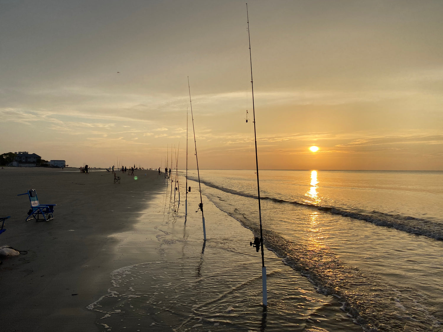 picture of a row of fishing poles in sand spikes along the beach with the sun rising in the background.